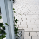 Podjazd i chodnik wyłożony jasnobeżową kostką brukową Semmelrock Lundo Bello, ułożoną w modularny wzór; widoczna biała konstrukcja z pnącym bluszczem oraz rabata z szarym grysem dekoracyjnym.